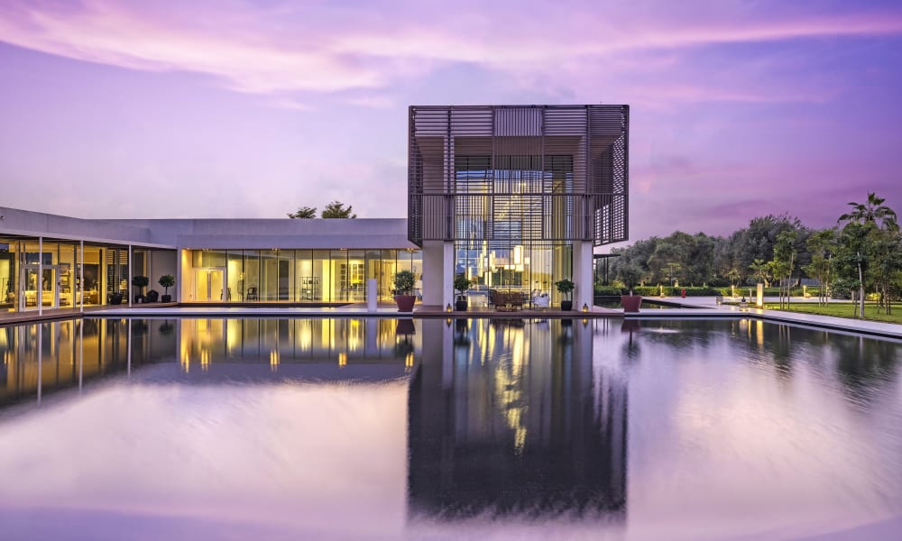 A modern resort with a central building reflected in a large, still pool, during sunset with purple and pink sky.