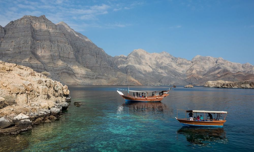 Zwei traditionelle Holzboote schwimmen auf klarem blauen Wasser vor rohen Bergen in Oman.