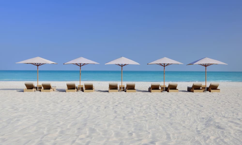 Sieben Strandstühle mit Sonnenschirmen entlang eines Sandstrandes mit Blick auf das ruhige Meer und einen klaren blauen Himmel.