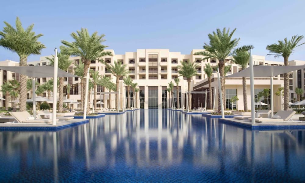 Luxuriöses Hotelkomplex mit großem Pool, umgeben von Palmen und modernen Gebäuden unter klarem blauem Himmel.