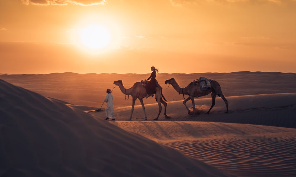 A person wearing white leads two camels across sand dunes at sunset, with another person riding one of the camels in a desert landscape.