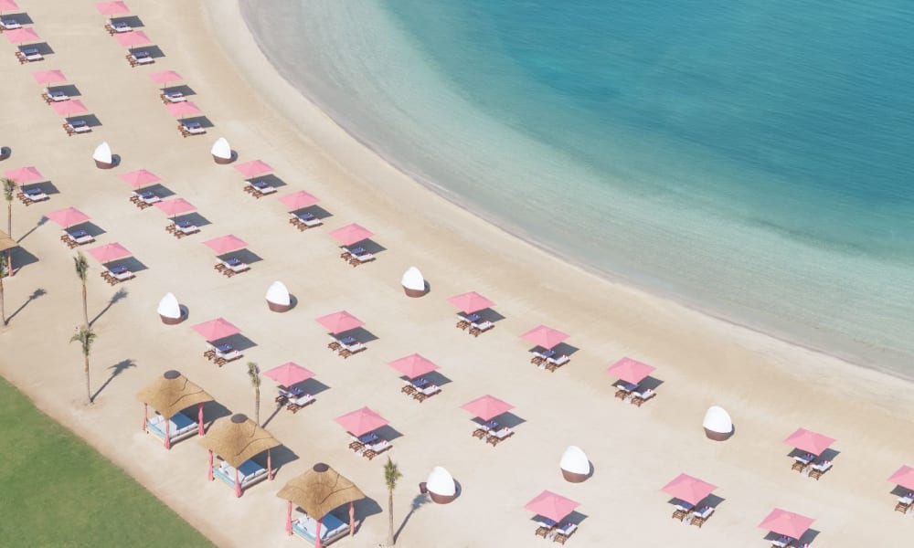 An aerial view of a beach with pink umbrellas and lounge chairs lined up along the shore, with small beach huts and palm trees nearby.