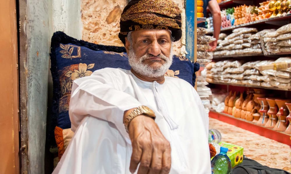 Ein älterer Mann mit weißem Bart, in traditioneller omanischer Kleidung und einer Mütze, sitzt vor einer strukturierten Wand mit einem Kissen hinter ihm in einem Markt.