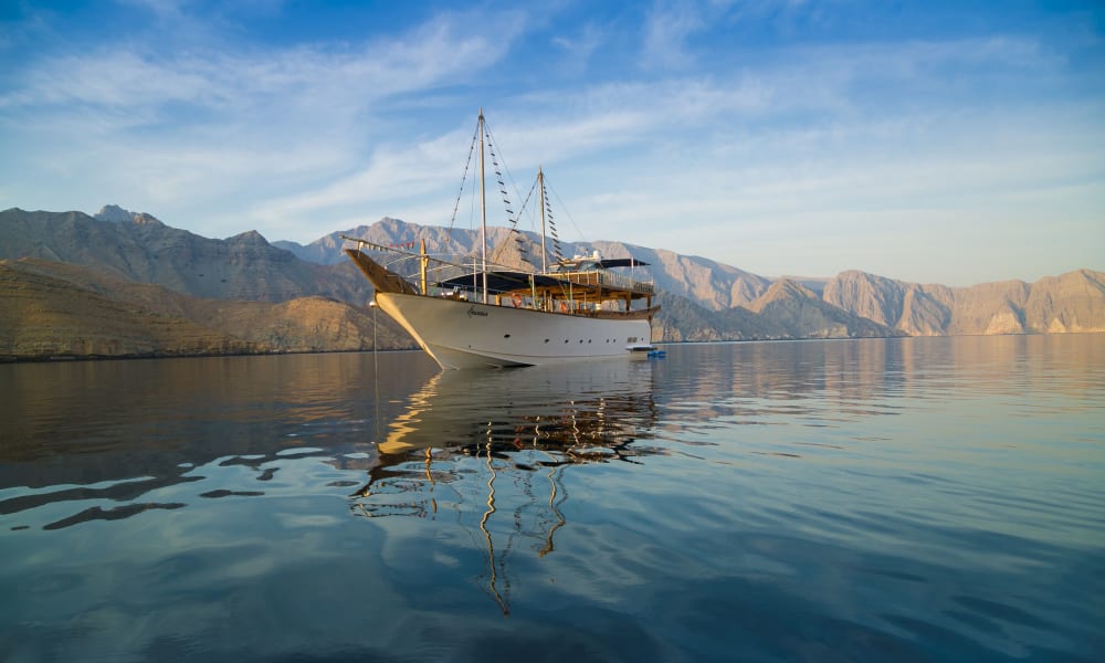 Ein weißes traditionelles Dhow-Boot treibt auf ruhigem Wasser vor einer bergigen Landschaft.