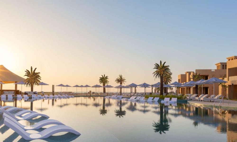 Ein luxuriöser Hotelpoolbereich bei Sonnenuntergang mit Liegestühlen, Sonnenschirmen und Palmen, die sich im Wasser spiegeln.