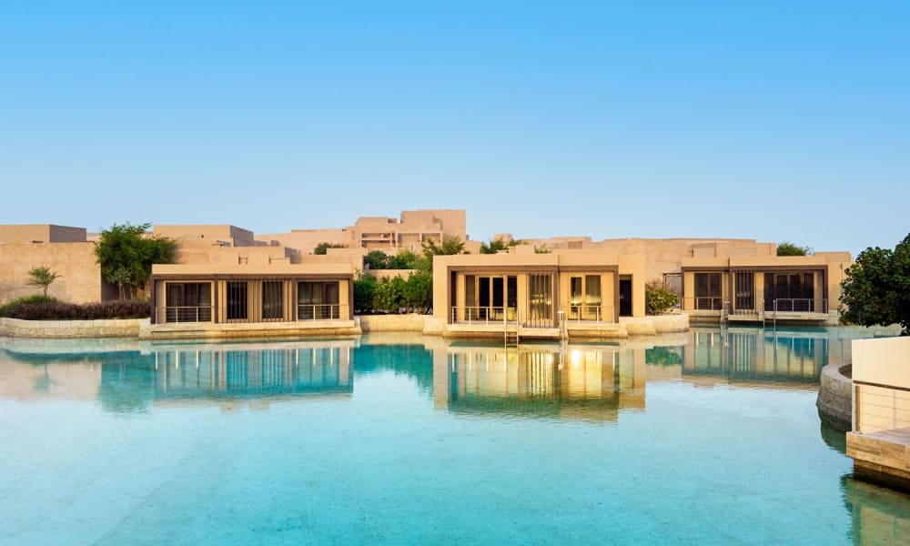 Luxury resort buildings reflected in a calm water body against a clear blue sky.