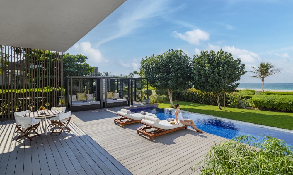 Zwei Frauen entspannen auf Sonnenliegen in der Nähe eines Pools an einem Frontresort mit üppigem Grün und dem Meer im Hintergrund.