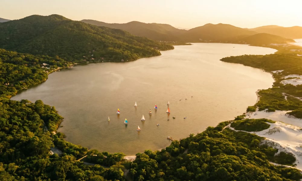 Luftaufnahme der Lagoa da Conceição in Florianópolis mit türkisem Wasser, weißen Sanddünen und Atlantikküste im goldenen Abendlicht