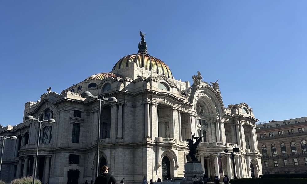 The Palacio de Bellas Artes in Mexico City under a clear blue sky, with a few people walking nearby on a paved area.