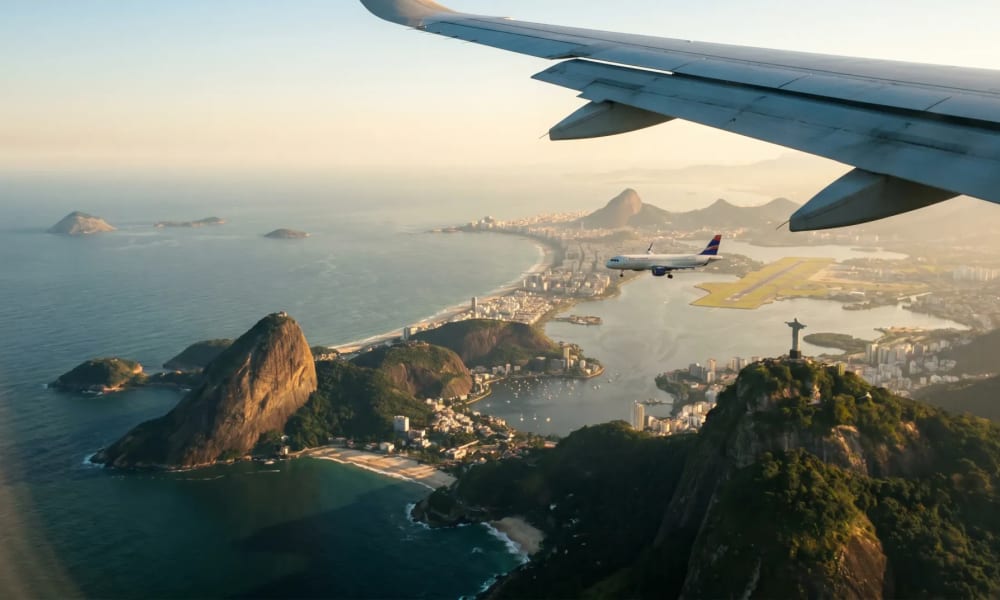 Luftaufnahme der Küste von Rio de Janeiro mit dem Zuckerhut, der Christus-Erlöser-Statue und einem Flugzeug, das nahe der Küste unter einem Flugzeugflügel fliegt.