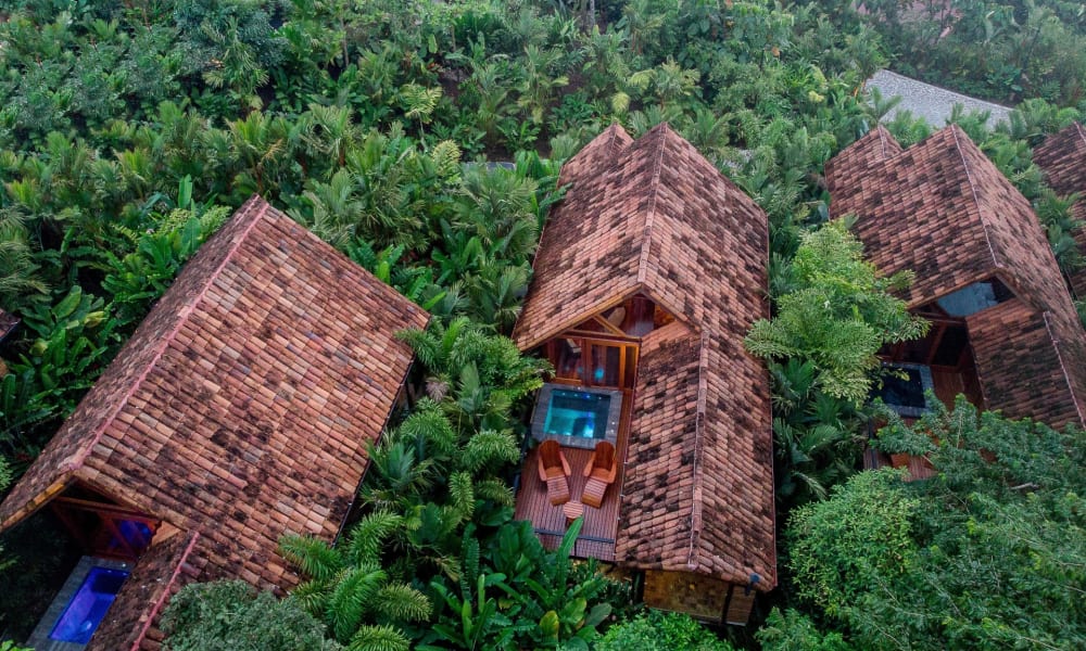 Drei traditionelle Holzhütten mit Satteldächern, umgeben von üppigem tropischem Grün, mit einer kleinen Holzterrasse und Whirlpool auf einer Veranda.