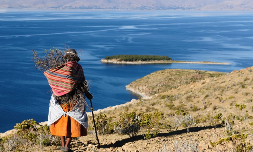 Ältere Frau in traditioneller Kleidung steht auf einem Hügel mit Blick auf den Titicacasee und Inseln im Hintergrund.