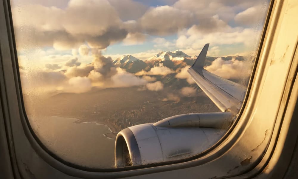 Blick aus dem Flugzeugfenster auf das Flugzeugflügel und Triebwerk mit schneebedeckten Bergen und Wolken in der Ferne bei Sonnenuntergang.