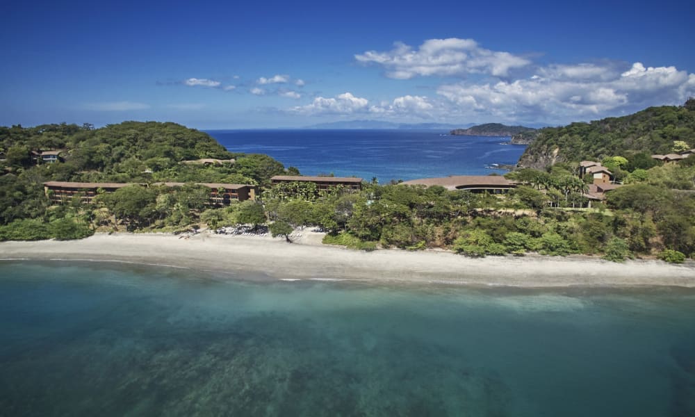 Luftaufnahme einer Küstenlandschaft mit Strand, üppigem Grün und Gebäuden zwischen den Bäumen unter einem wolkigen Himmel.
