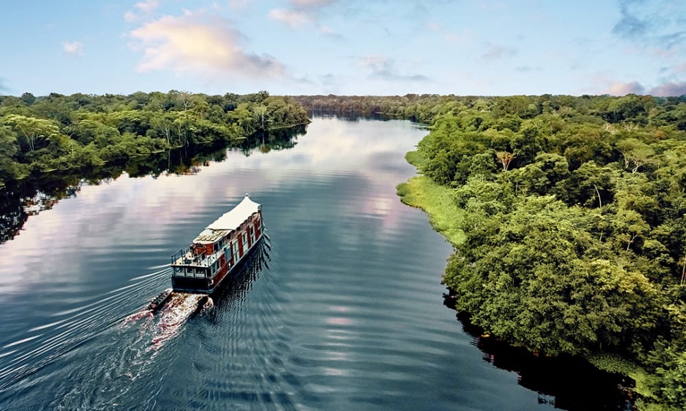 Ein Boot fährt auf einem breiten Fluss durch dicht bewachsenen grünen Regenwald unter einem teils bewölkten Himmel.