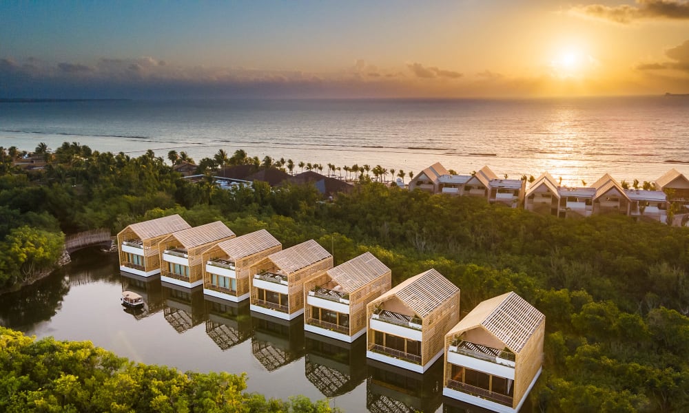 Sonnenuntergang über einer Reihe von modernen hölzernen Strandvillen mit Dachterrassen, die sich in einer Lagune spiegeln, umgeben von dichtem Grün und mit Blick auf das Meer.
