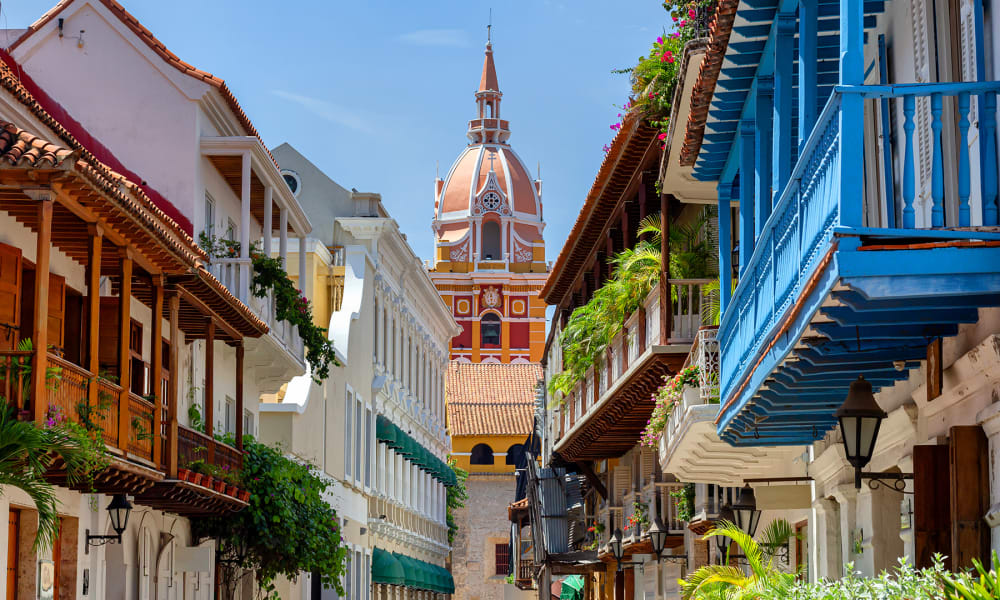 Bunte koloniale Gebäude mit Holzbalkonen in einer Straße von Cartagena, Kolumbien, mit der Kuppel der Kathedrale von Cartagena im Hintergrund unter klarem blauem Himmel.