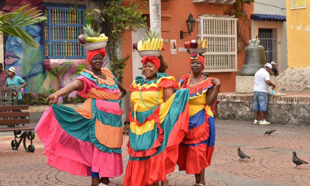 Three women in colorful traditional dresses standing on a cobblestone street in Cartagena de Indias, Colombia, with fruit baskets balanced on their heads and vibrant street art and colonial buildings in the background.