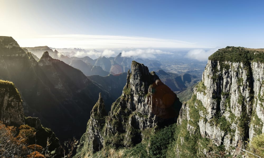 Rocky mountain cliffs covered with some vegetation under a bright, clear sky with distant misty peaks and valleys.
