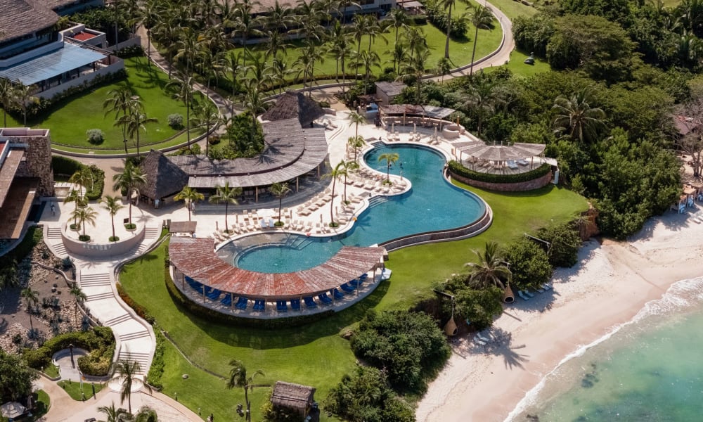 Luftaufnahme eines Resort-Anwesens mit einem einzigartig geformten Pool mit Liegestühlen, umgeben von Palmen, grünen Rasenflächen und angrenzend an einen Sandstrand mit klarem Meerwasser.