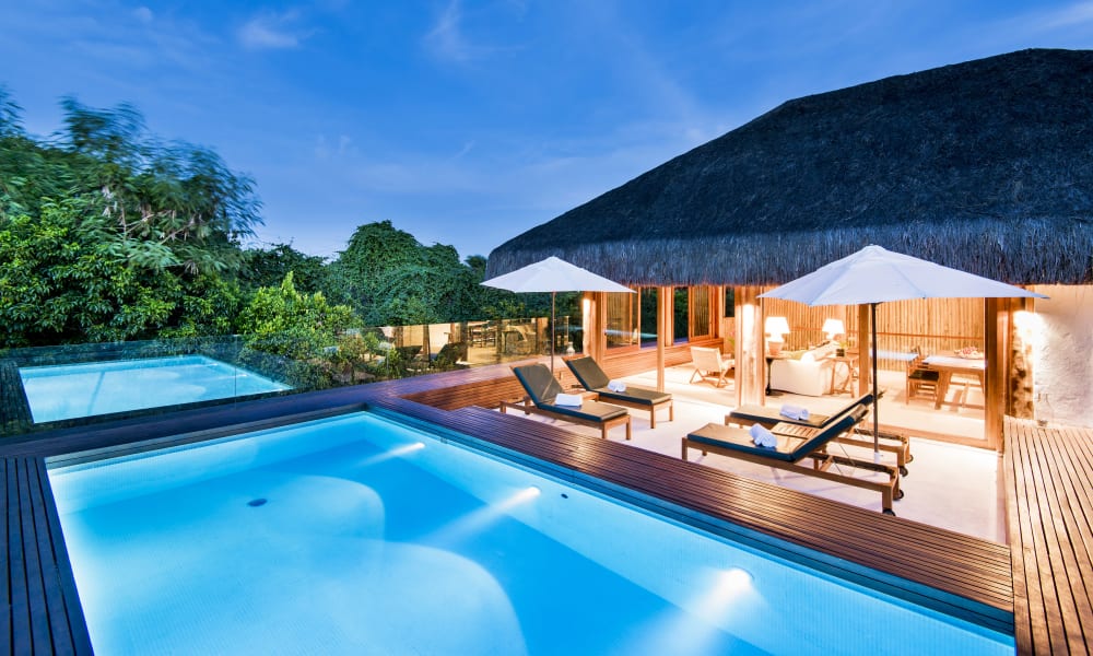 Ein luxuriöser Außenpoolbereich eines tropischen Resorts mit Sonnenliegen und Sonnenschirmen, neben einer Strohdach-Villa.