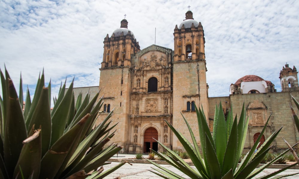 Historische Steinkirche mit zwei Türmen in Oaxaca, eingefasst von Agavenpflanzen unter einem teilweise bewölkten Himmel.