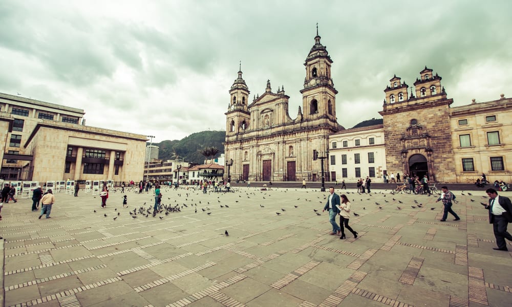 Großer öffentlicher Platz in Bogotá, Kolumbien, mit historischen Kolonialgebäuden und einer Kathedrale unter bewölktem Himmel, mit Spaziergängern und verstreuten Tauben.
