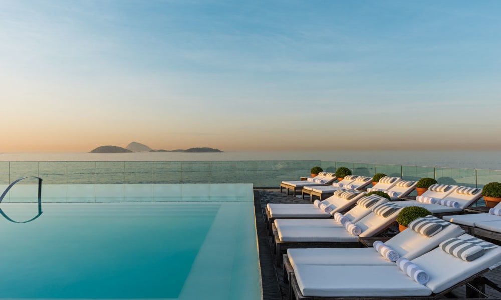 Infinity-Pool mit weißen Liegestühlen und eingerollten Handtüchern auf der rechten Seite, Blick auf ruhiges Meerwasser und entfernte Inseln unter klarem Himmel bei Sonnenuntergang.