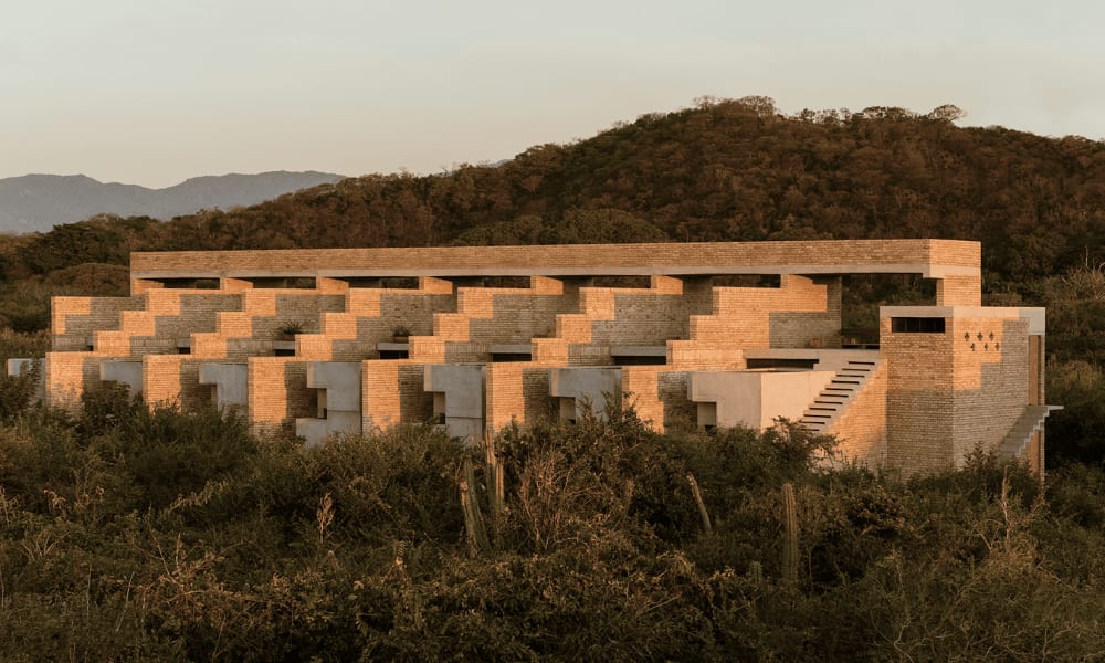 Langes, beiges Steingebäude mit gestufter architektonischer Gestaltung in einer trockenen, mit Gestrüpp bedeckten Landschaft, umgeben von Hügeln unter klarem Himmel.