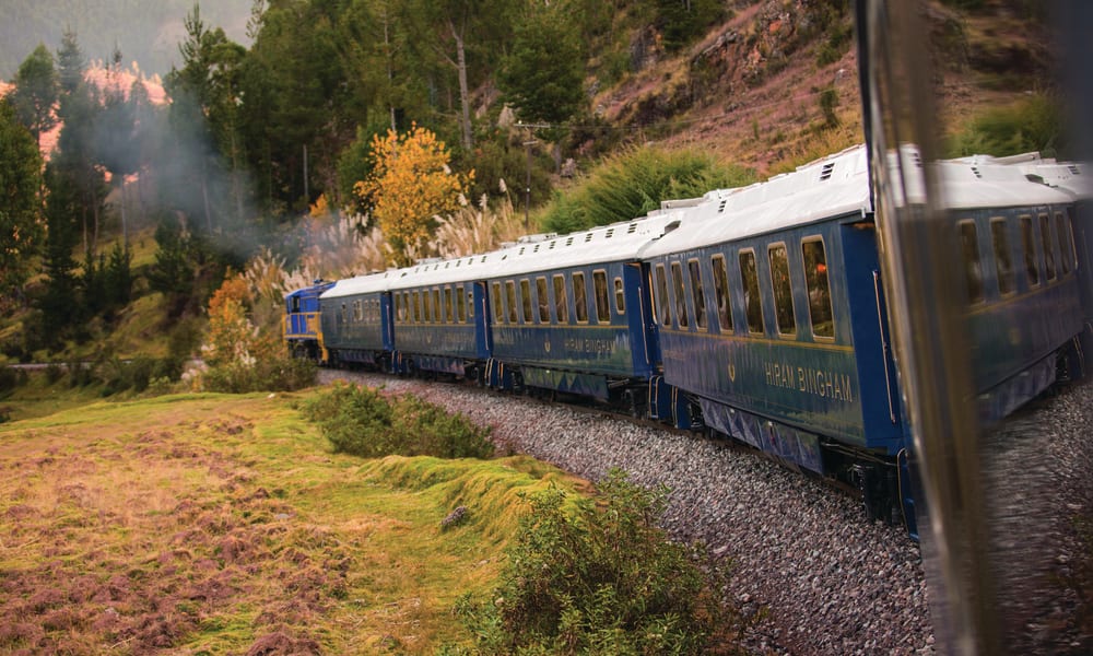 Blue Hiram Bingham train moving along a track surrounded by green vegetation and trees with a mountainous backdrop.