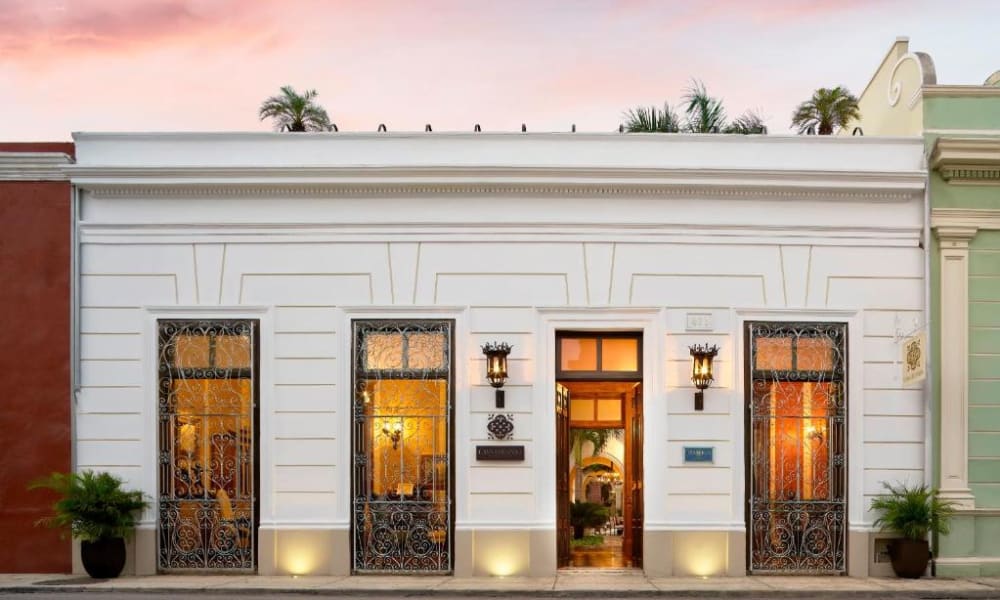 Elegante weiße Fassade des Casa Lecanda Boutique Hotels mit beleuchteten Fenstern und Eingang, verziert mit Schmiedeeisendetails und Blumentöpfen auf beiden Seiten unter einem rosa gefärbten Abendhimmel.