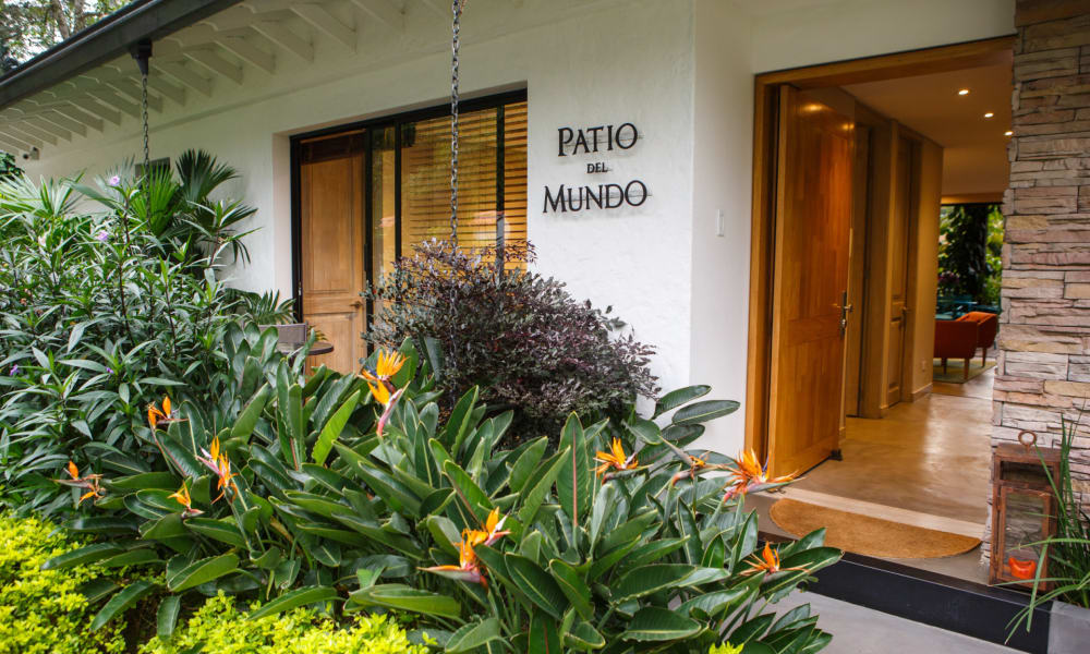 Der Eingang zum Patio del Mundo in Medellín, Kolumbien, ist umgeben von üppigem Grün mit bunten blühenden Pflanzen und einer modernen Holztür.