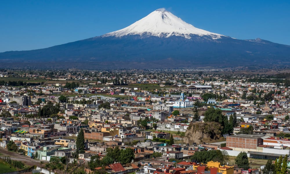 Luftaufnahme der Stadt Puebla mit zahlreichen Gebäuden und Straßen im Vordergrund, vor dem Hintergrund eines großen schneebedeckten Vulkans unter klarem blauem Himmel.