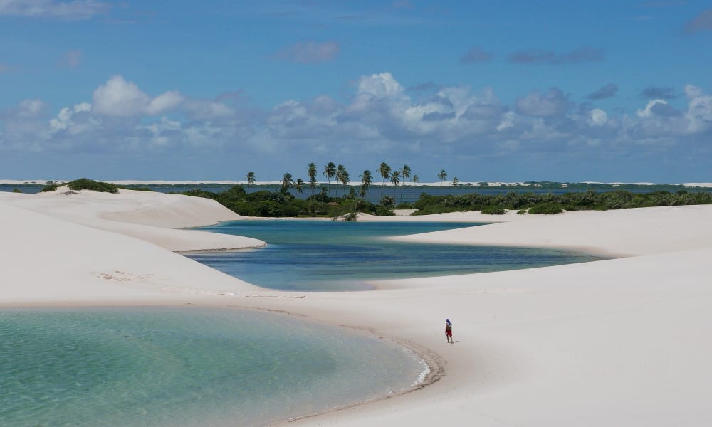 A person walking on white sand dunes near clear blue lagoons with palm trees and green vegetation in the background under a partly cloudy sky.
