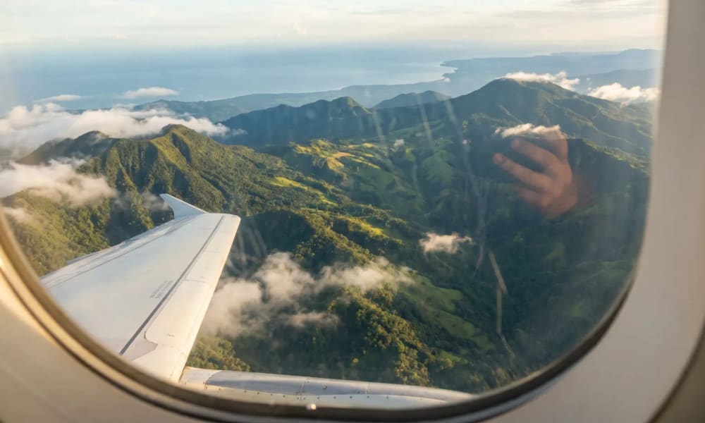 Blick aus dem Flugzeugfenster auf grüne Berge und Wolken über einer Küstenlandschaft bei Tageslicht.