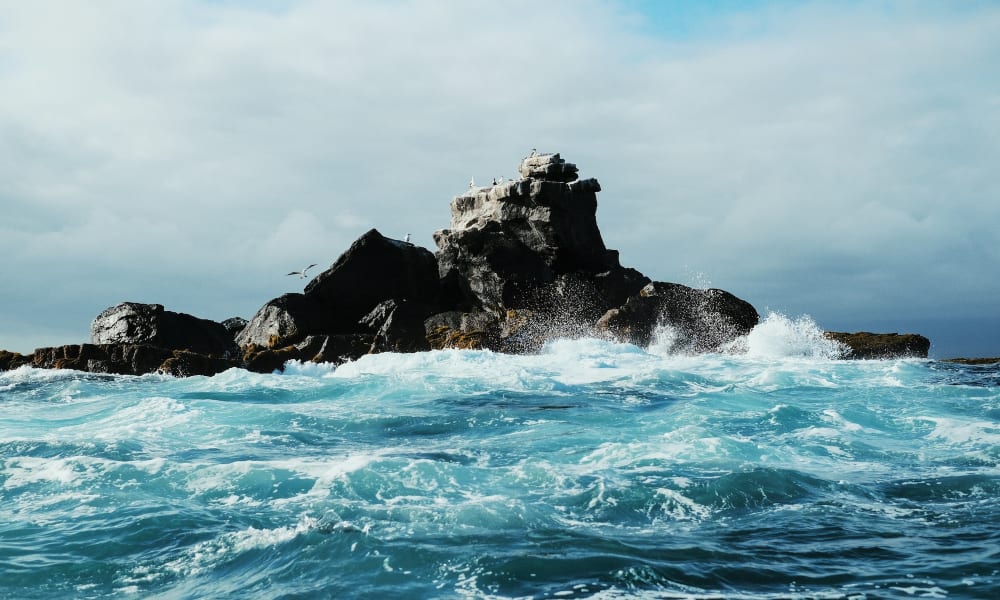 Rough ocean waves crash against dark volcanic rocks in the sea.