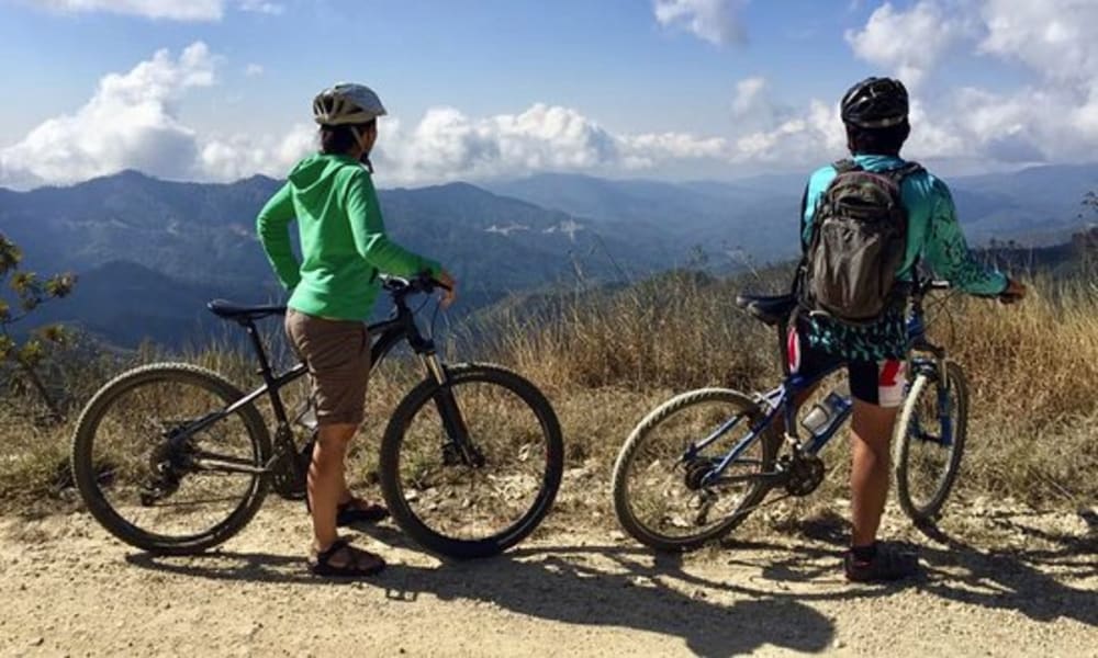Zwei Mountainbiker stehen auf einem Schotterweg mit Blick auf eine malerische Berglandschaft und teilweise bewölkten Himmel am Tag.