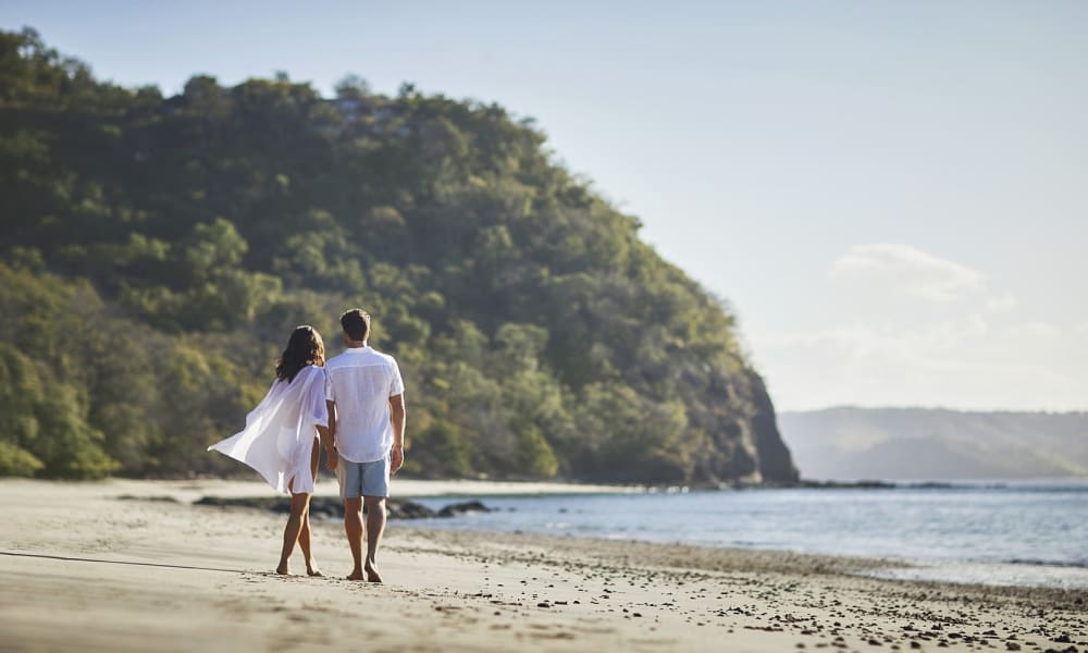 Ein Paar geht Hand in Hand am Sandstrand entlang mit üppig grünen Hügeln und ruhigem Meer im Hintergrund in Papagayo, Costa Rica.