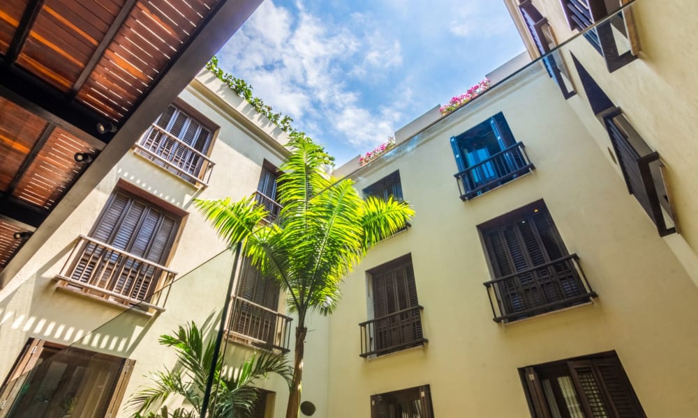 View from the courtyard of a hotel looking up at the sky, surrounded by cream-colored walls with black window shutters and balconies.