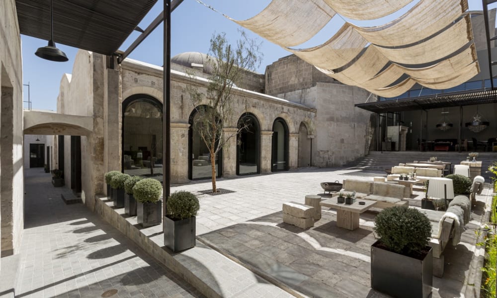 An outdoor lounge area with modern seating, potted plants, and shaded canopy in a spacious courtyard.