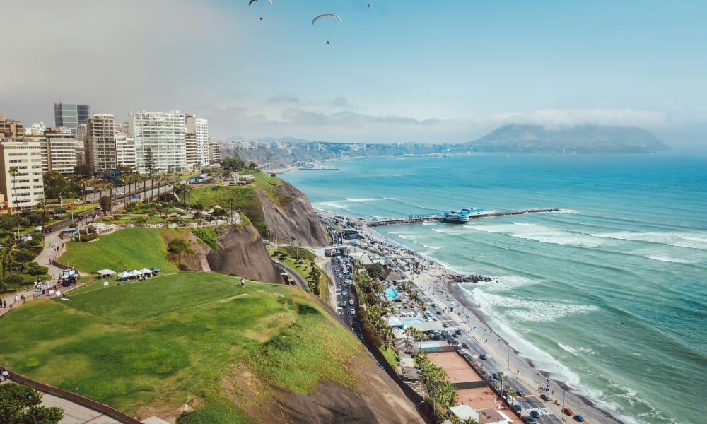 Stadtansicht entlang der Küste von Lima in Peru mit Hochhäusern, Klippen, grünen Parkflächen, einem Pier, der ins Meer ragt, und drei Gleitschirmfliegern am Himmel.