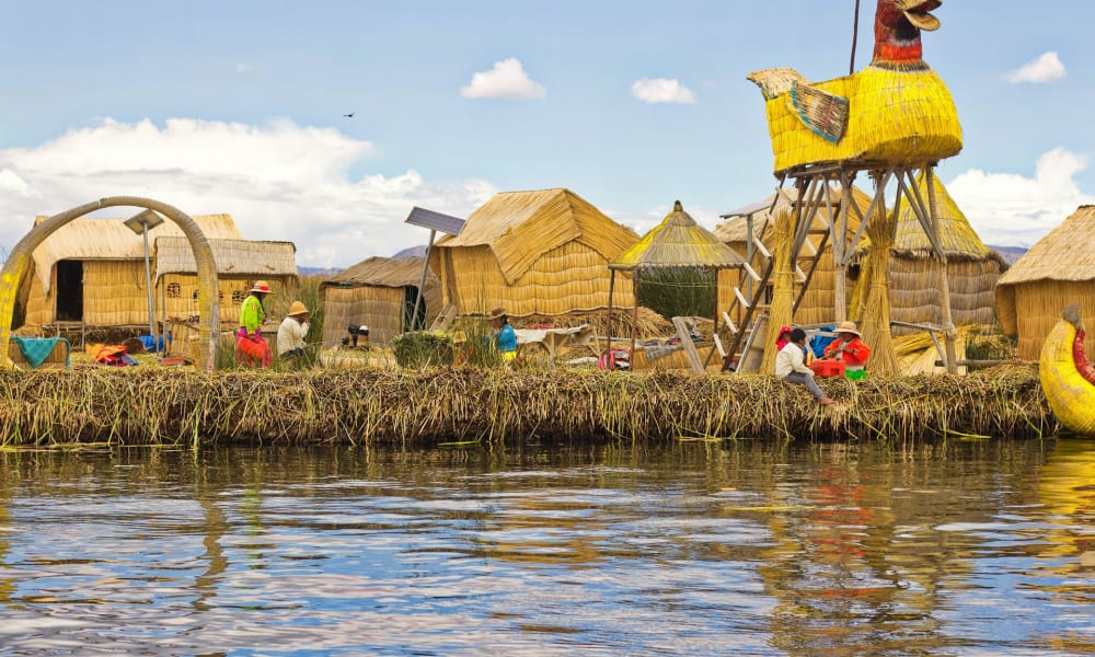 Ein Dorf am See mit traditionellen Strohdächern und großen gelben vogelförmigen Strukturen aus Schilf, mit Menschen, die nahe am Wasser täglichen Aktivitäten nachgehen, unter einem teils bewölkten Himmel.