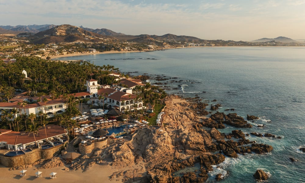 Luftaufnahme des Resorts One&Only Palmilla am Morgen mit Gebäuden, Schwimmbad, Palmen, felsiger Küste und ruhigem Meer unter leicht bewölktem Himmel.