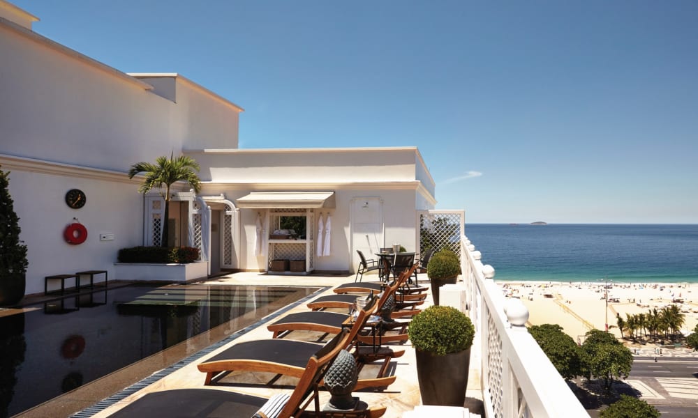 A luxurious hotel terrace overlooking the beach in Rio de Janeiro with sun loungers lined up and potted plants.