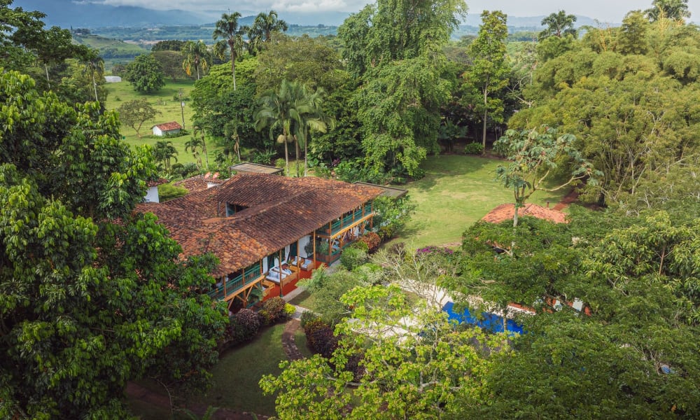 Luftaufnahme der Hacienda Bambusa, umgeben von dichtem grünem Baumbestand und Vegetation mit einem klaren Hof und bewölktem Himmel im Hintergrund.