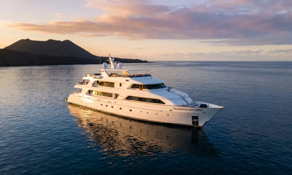 Eine weiße Luxusyacht namens 'Galapagos Explorer' fährt bei Sonnenuntergang auf ruhigem Ozeanwasser nahe vulkanischer Inseln.