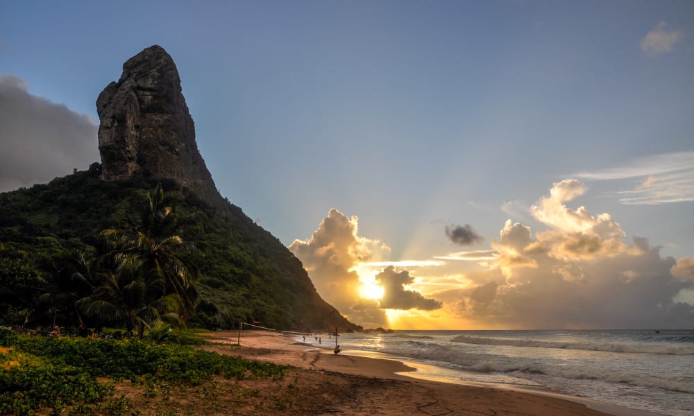 A scenic beach at sunset with a towering rocky hill on the left and the ocean on the right, with rays of sunlight breaking through clouds.