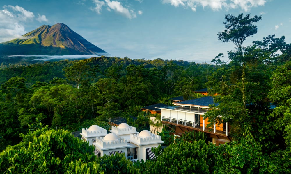 Luftaufnahme des Nayara Springs Resorts, umgeben von dichtem grünen Wald mit einem Vulkangebirge im Hintergrund unter einem teils bewölkten Himmel.