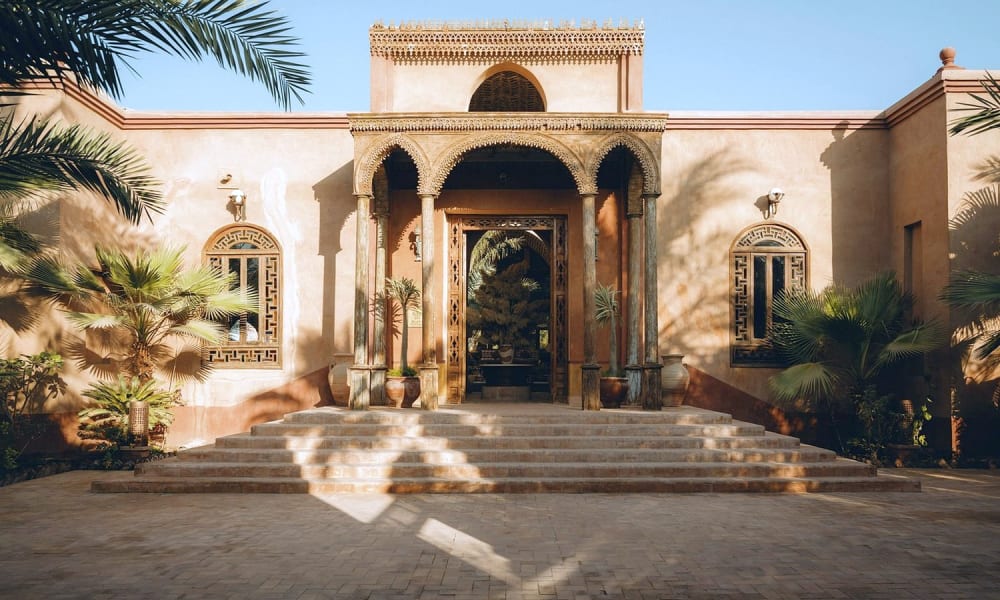 Großer Eingang zum Al Moudira Hotel mit traditioneller Architektur, Palmen und Steinstufen, die zur Tür führen.