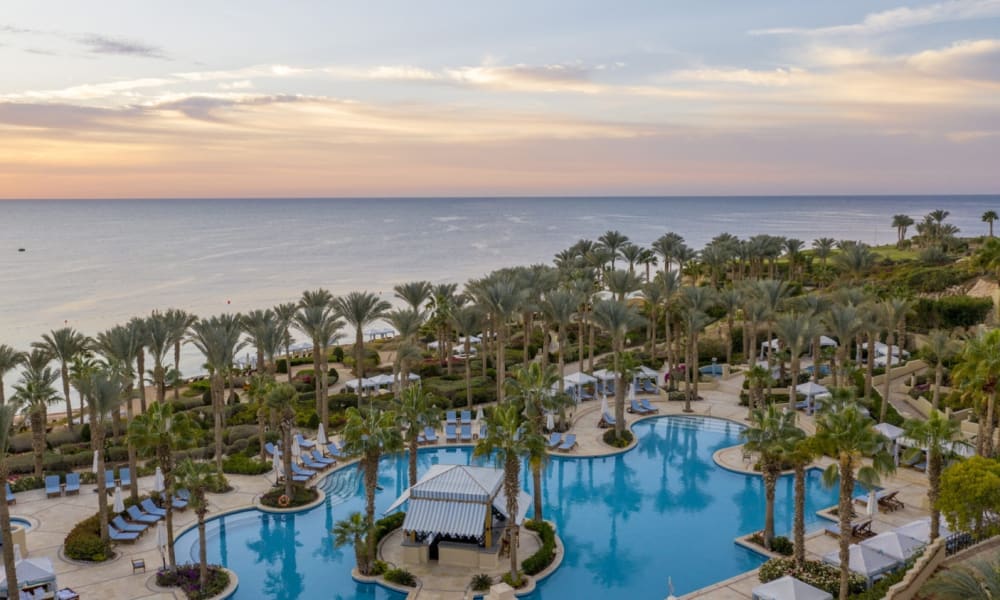 Eine luxuriöse Resort-Poolanlage mit zahlreichen Palmen, Sonnenliegen und einem Pavillon, Blick auf den Strand und das Meer bei Sonnenuntergang.